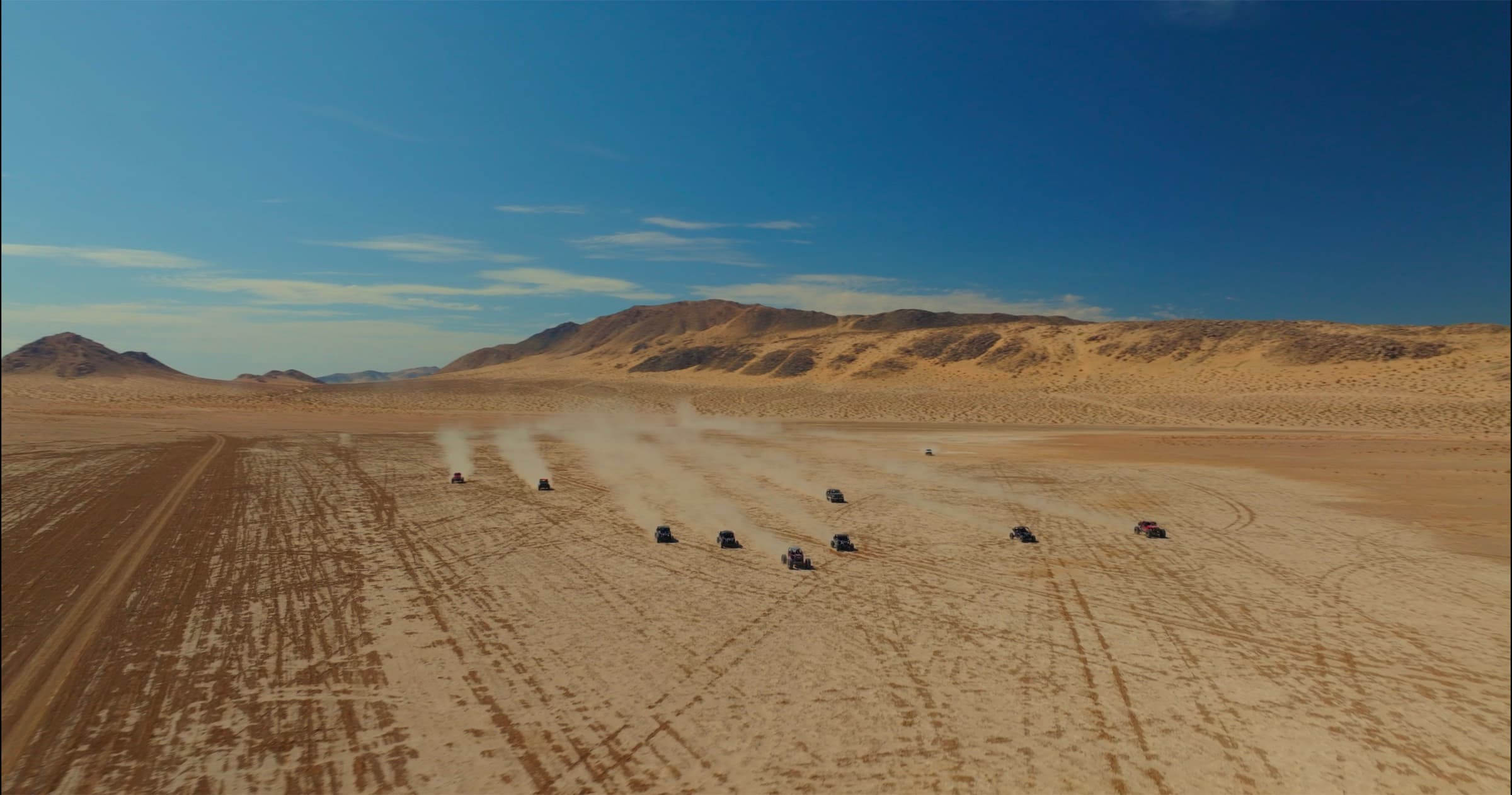 A formation of off-road rigs racing across the dry lakebed, kicking up dust